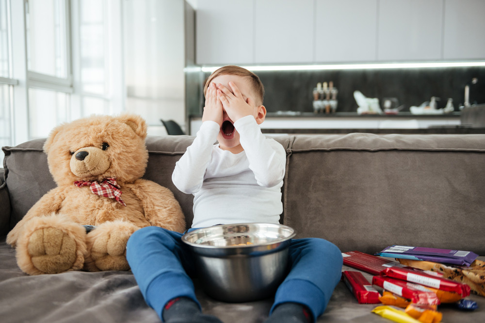 Photo of scared little boy on sofa with teddy bear at home watching TV while eating chips.