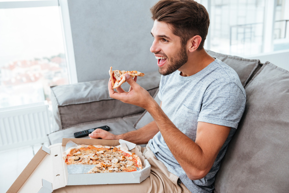 Picture Of Cheerful Hangry Young Man Eating Pizza While Sitting On Sofa And Watching Tv Royalty 