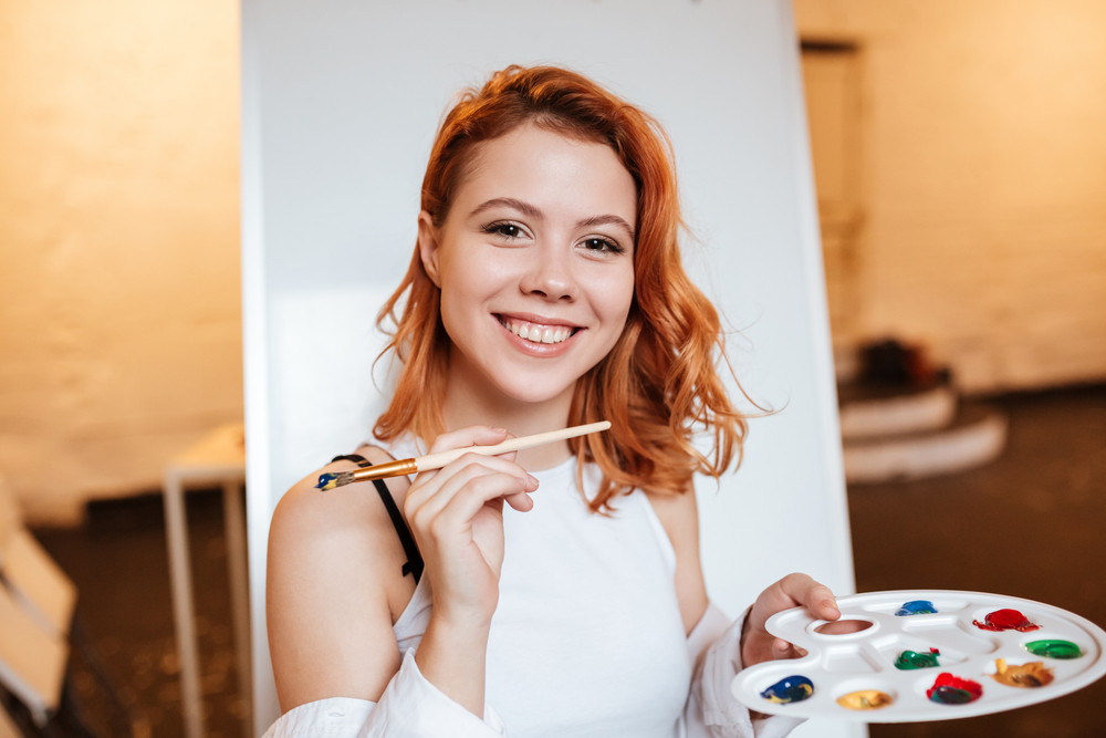 Picture of happy redhead young woman painter standing over blank canvas in artist workshop. Look at camera while holding palette and paintbrush.