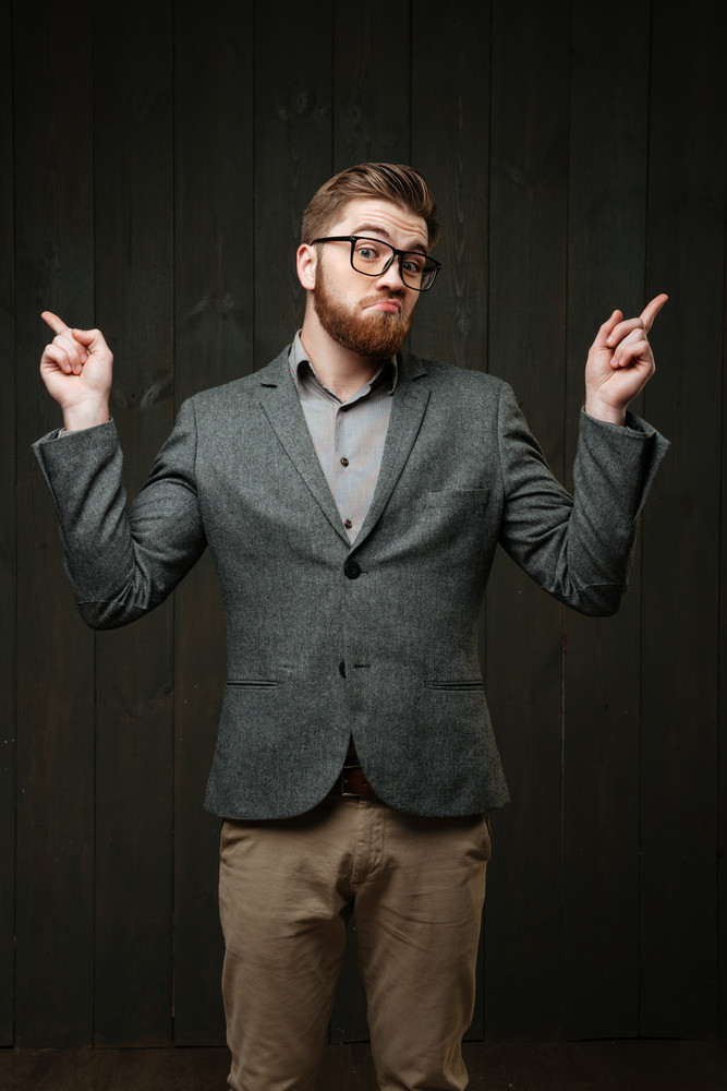Portrait of a puzzled bearded man in eyeglasses and suit pointing fingers away isolated on the black wooden background