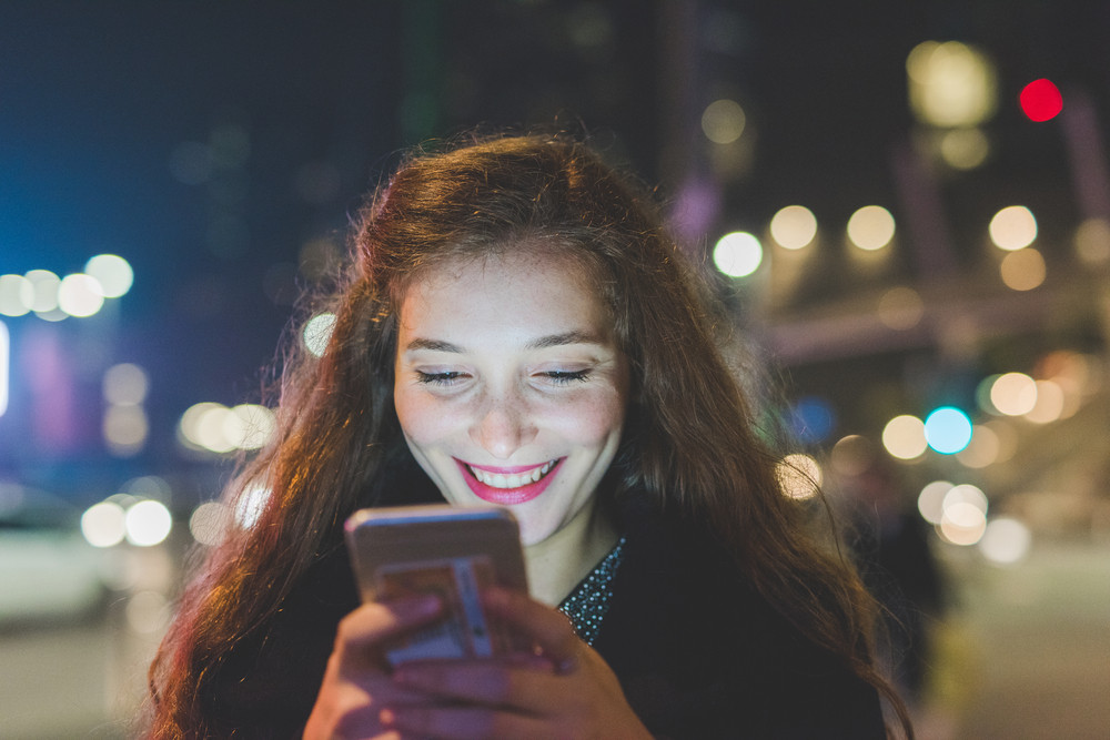 Portrait of young beautiful caucasian long brown hair woman using smart phone hand hold outdoor in the city night, smiling, face illuminated screenlight - social network, technology, comunication concept