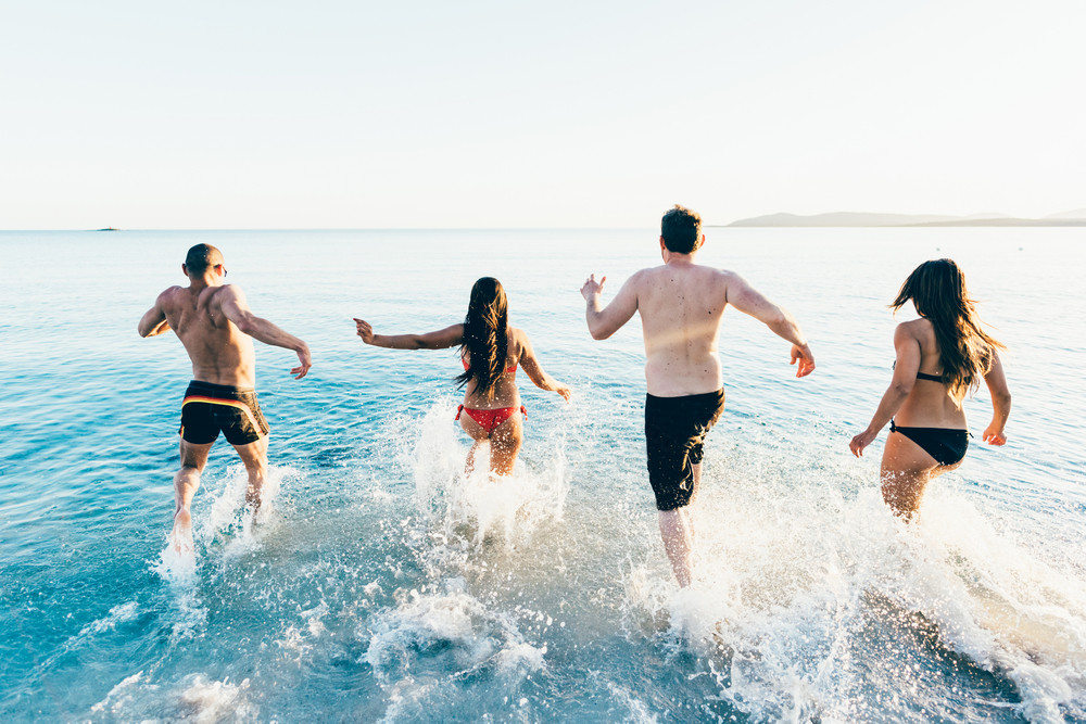 Rear view of a group of young multiethnic friends women and men running on the beach to the sea in summertime - happiness, summer, friendship concept
