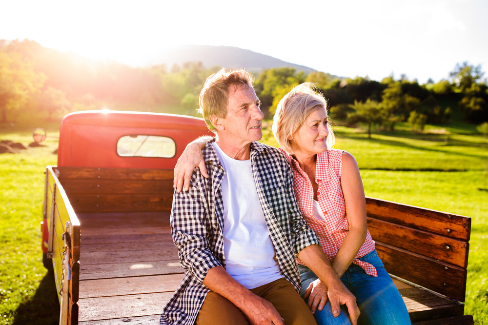 Senior couple sitting in back of vintage red pickup truck, hugging, green sunny nature