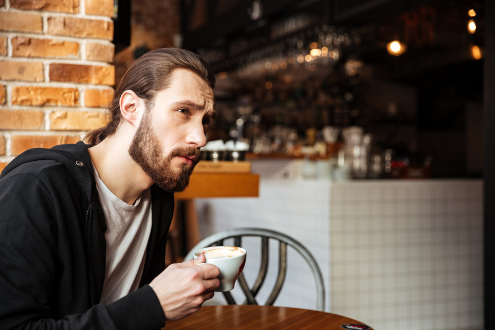 Side View Of Serious Bearded Man Sitting By The Table In Cafe With