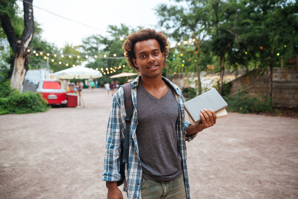 Smiling african young man with backpack standing and holding books outdoors