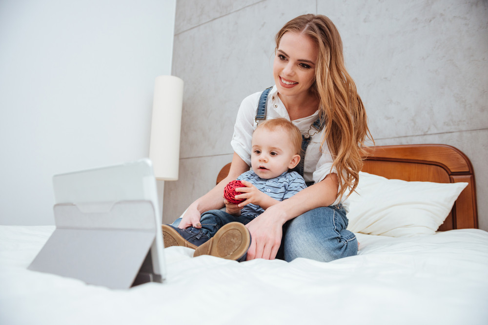Smiling young woman sitting with her son on bed and watching cartoons on tablet