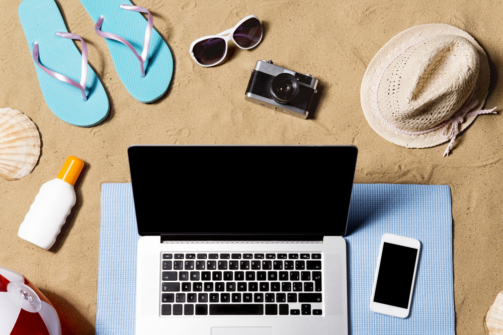 Summer vacation composition with laptop, smart phone, a pair of blue flip flop sandals, hat, sunglasses, sun screen and other stuff on a beach. Sand background, studio shot, flat lay, copy space.