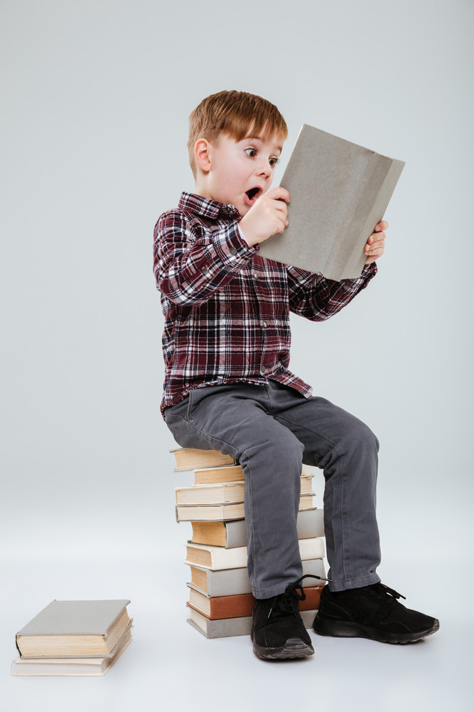 Vertical image of young surprised boy in shirt reading book and sitting on books. Isolated gray background