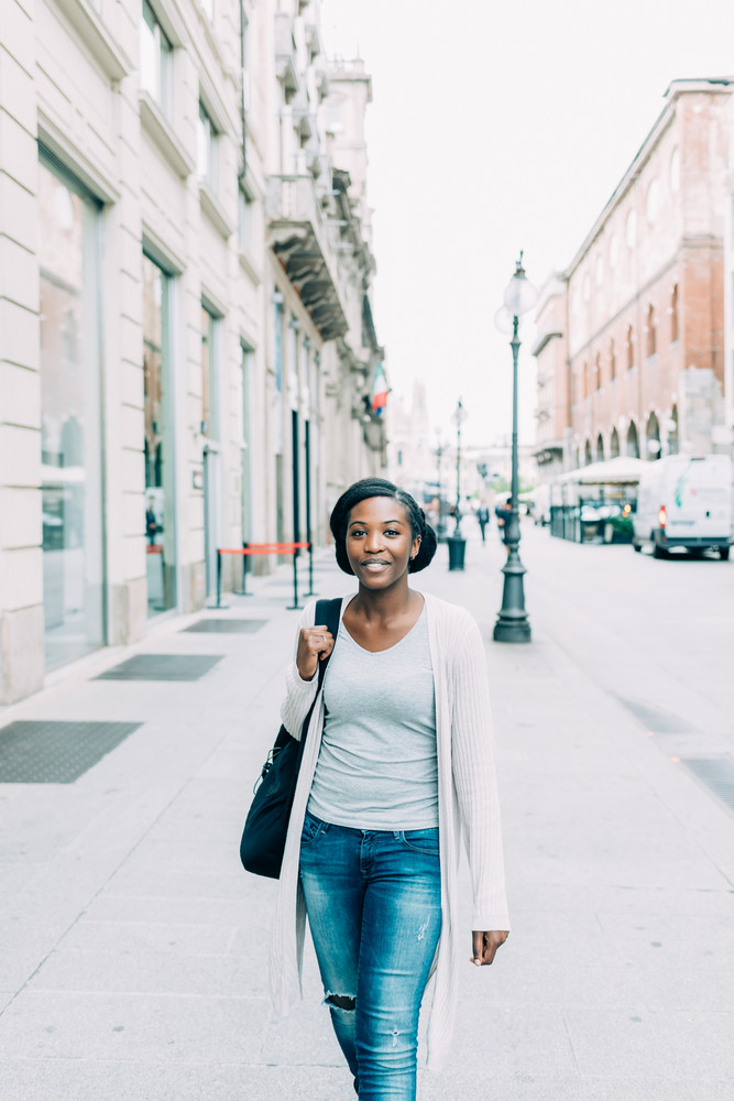 Young beautiful black woman outdoor in the city, looking at camera smiling wearing back pack - happiness, carefree, serene concept