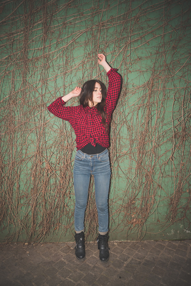 Young beautiful brunette long hair girl posing in front of a green wall touching her hair dressed with checked red shirt, blue jeans and black shoes. Light obtained due to the use of a flash