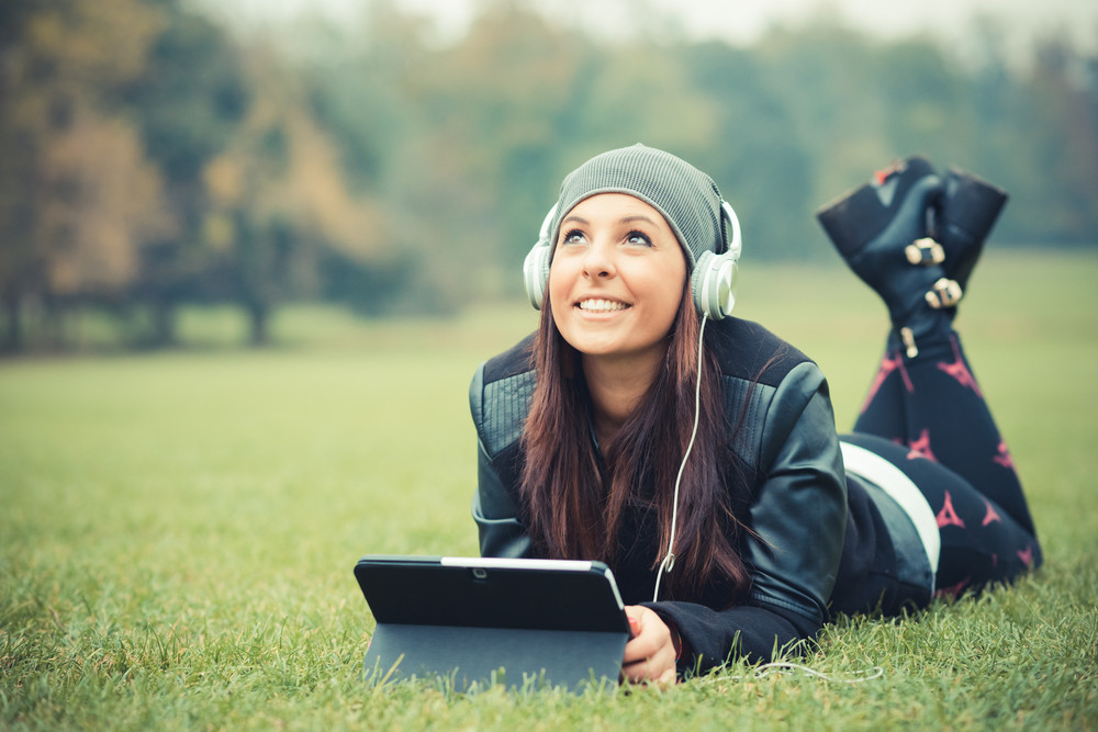young beautiful brunette straight hair woman in the park during autumn season - listening music with headphones and using tablet device connecting internet wireless