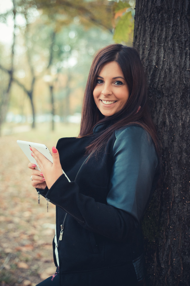 young beautiful brunette straight hair woman in the park during autumn season - using tablet technological device connecting web online wireless