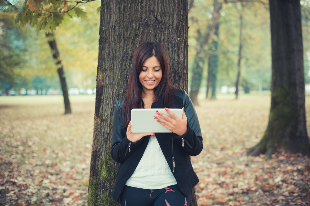young beautiful brunette straight hair woman in the park during autumn season - using tablet technological device connecting web online wireless