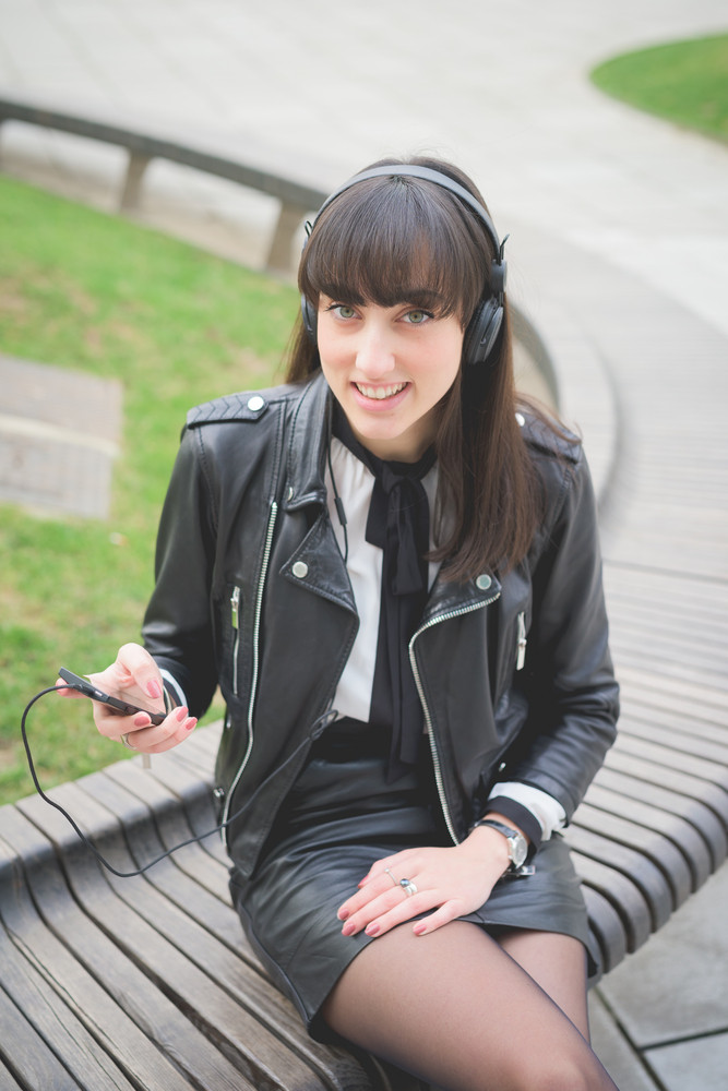 Young beautiful caucasian brown hair woman seated on a bench in a city park listening to the music looking in camera - relax, music concept