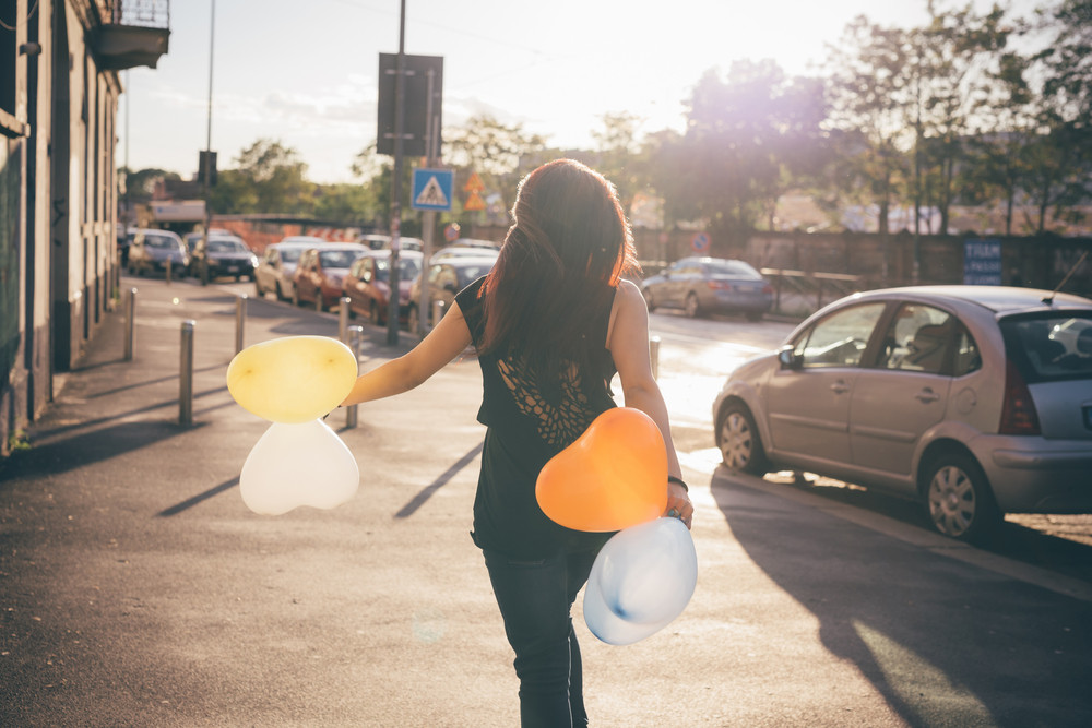 Young beautiful reddish brown hair caucasian woman viewed from back walking through the street playing with baloon in shape of heart - carefreeness, childhood, youth concept