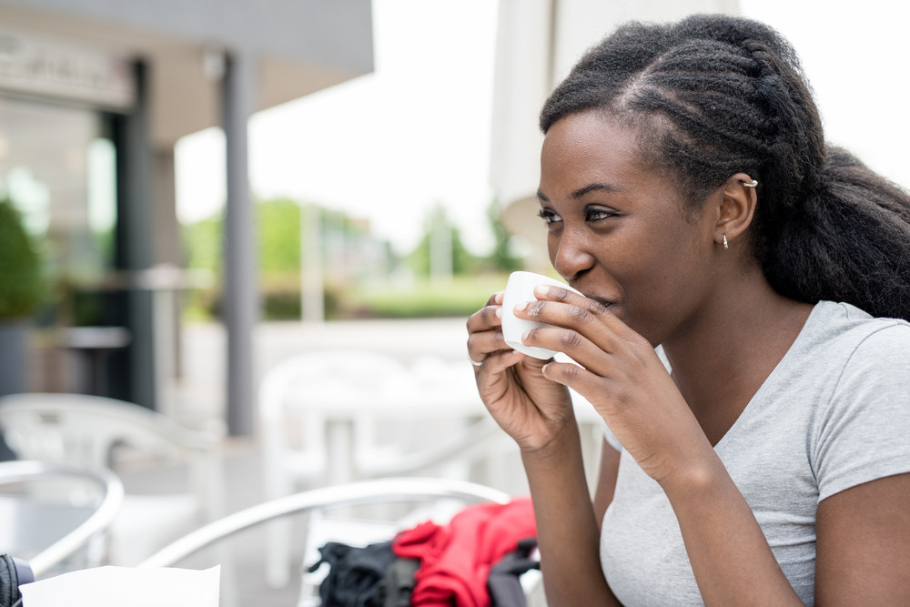 Young black woman having a cup of coffee outdoor in a bar, overlooking smiling - relax, break, happiness concept