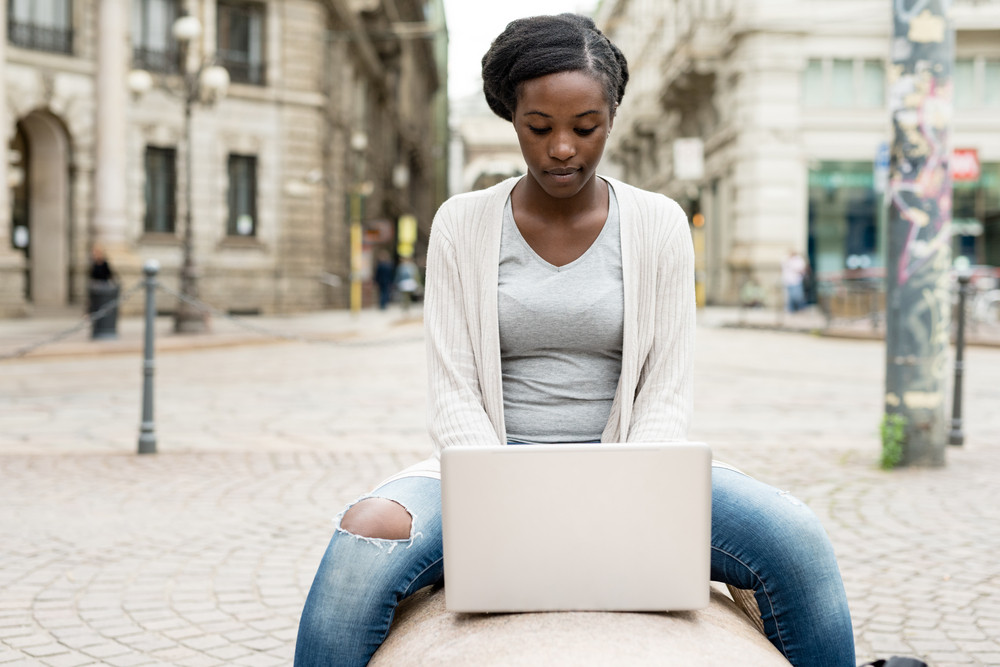 Young black woman using computer outdoor in the city sitting on a bench tapping the keyboard - business, student, technology concept
