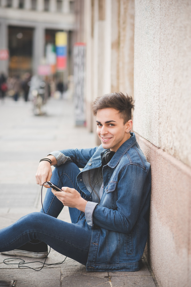 young handsome alternative dark model man sitting on the sidewalk with smart phone hand hold and headphones, looking at camera, smiling - happiness, carefree, thoughtless concept