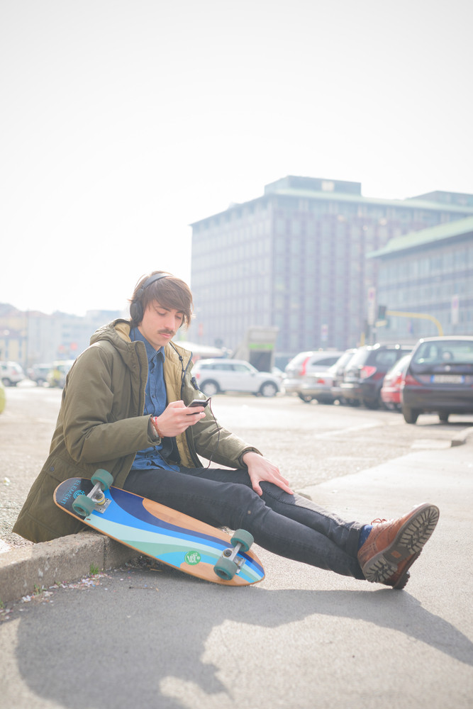 Young handsome caucasian man skater with moustache seated on a sidewalk listening to the music - relax, music concept - dressed with green coat and black jeans