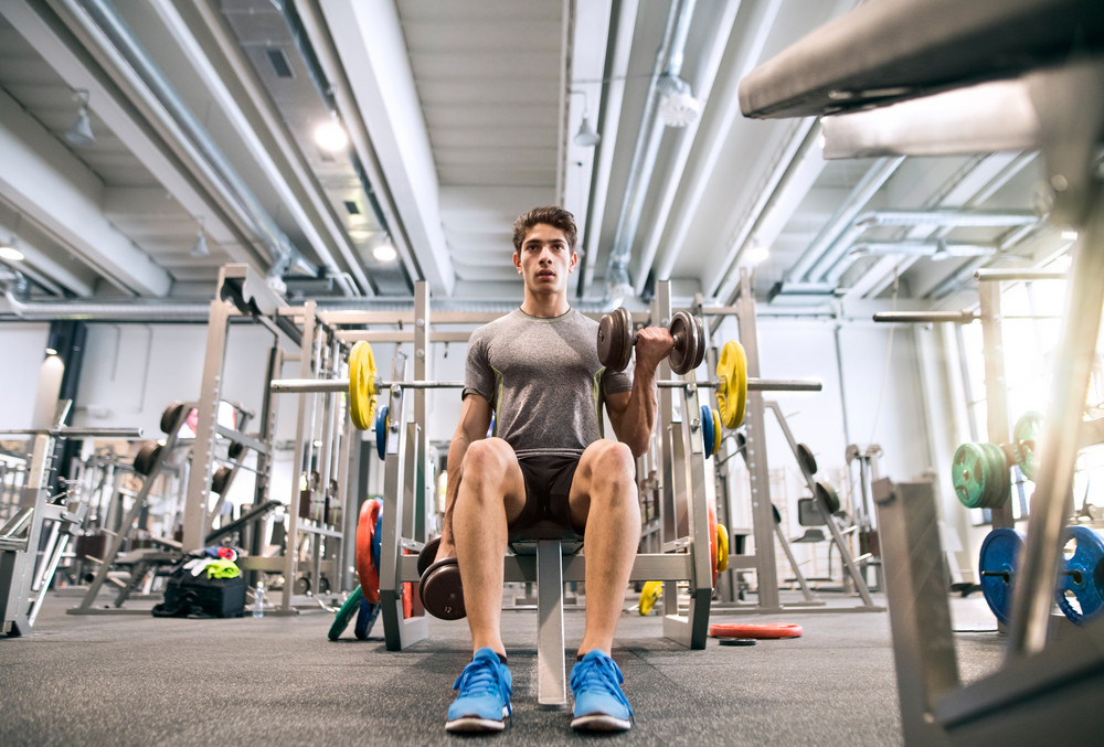 Young hispanic fitness man in gym sitting on bench, working out with weights