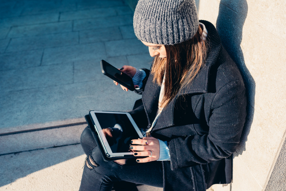 young woman tapping the screen of a tablet leaning on her knee and holding a smartphone on the other hand - multitasking, technology, social network concept