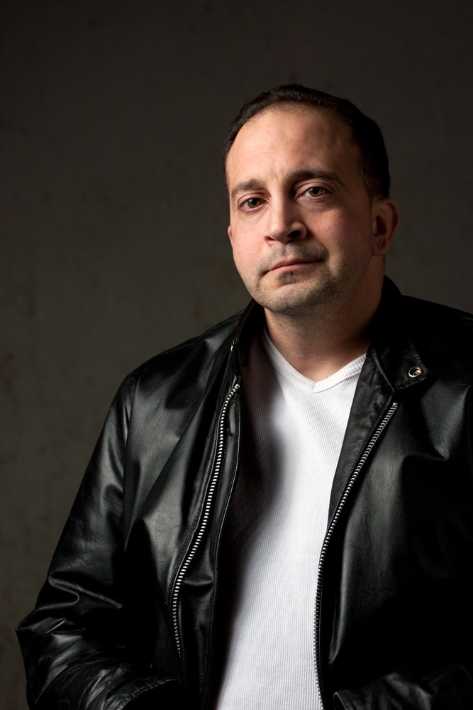 Portrait of a serious middle aged man in his upper 30s wearing a leather jacket in front of a grungy background.