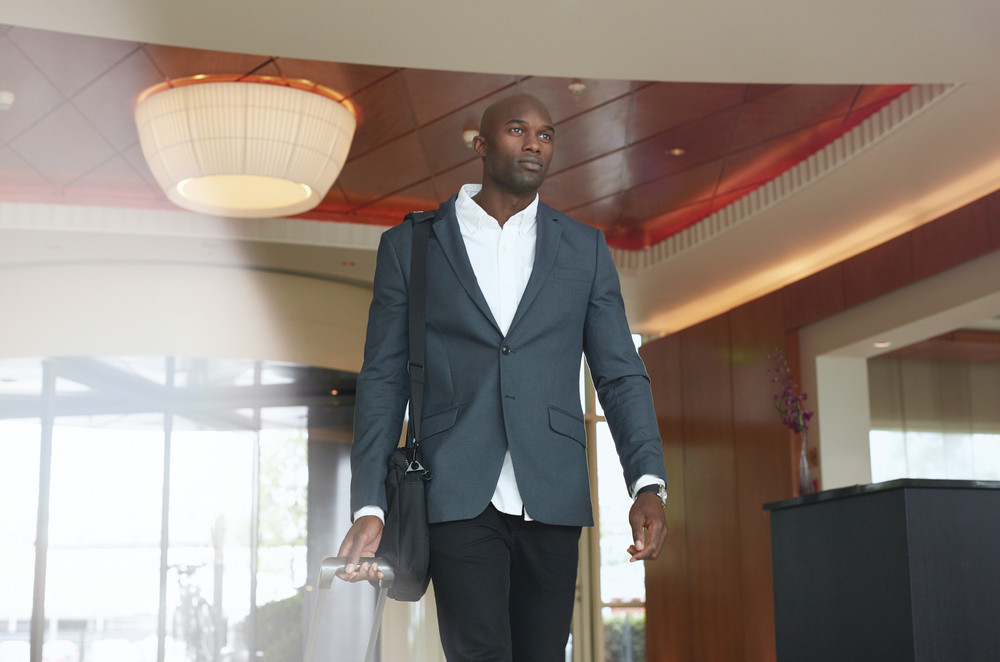 Portrait of young businessman with luggage in hotel lobby. African business executive walking in hotel hallway with his baggage.