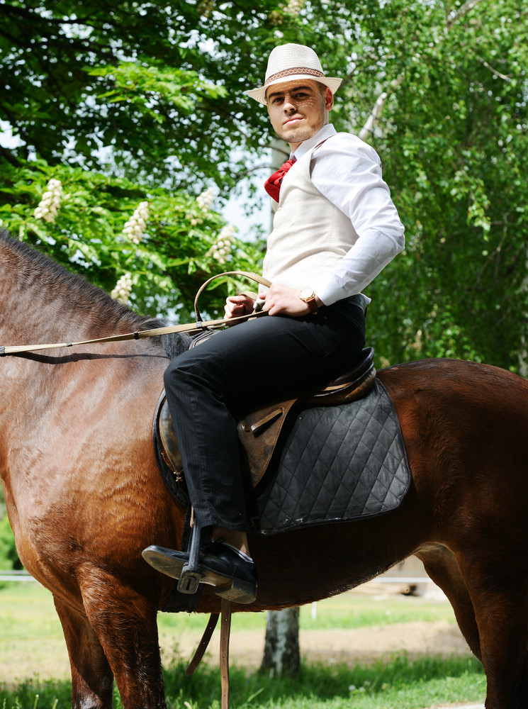 Young stylish man with tie and hat riding a horse on countryside