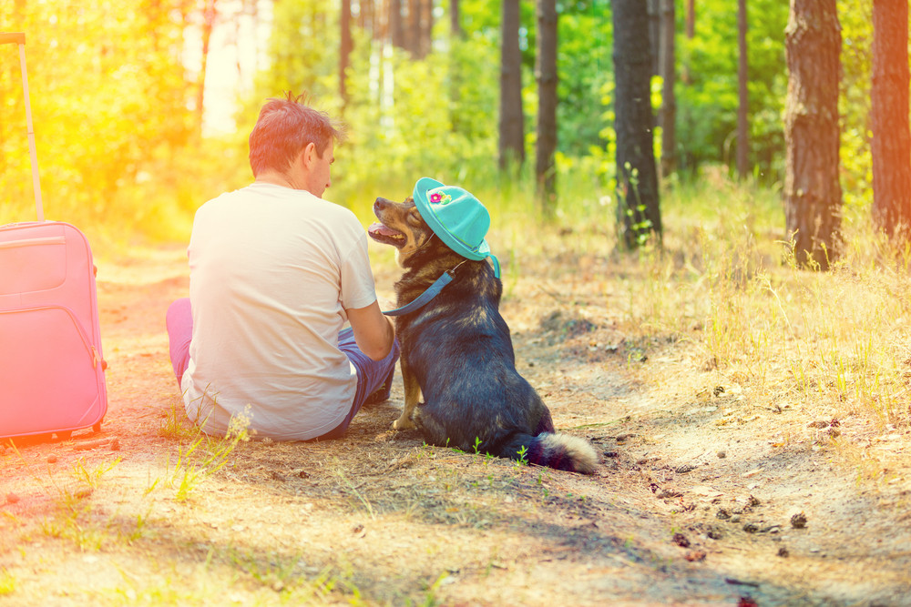 A man is sitting with a dog on a path in the forest. The dog is wearing a sun hat