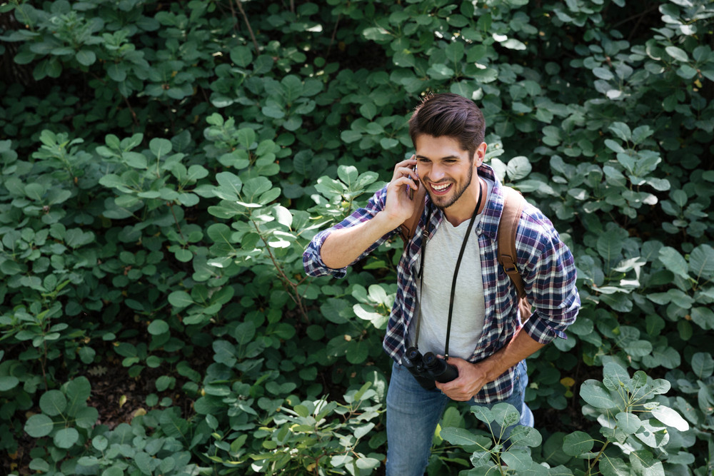 From above image of young guy in forest in the midst of plants. so smiling man. talking on the phone. guy with backpack and binoculars