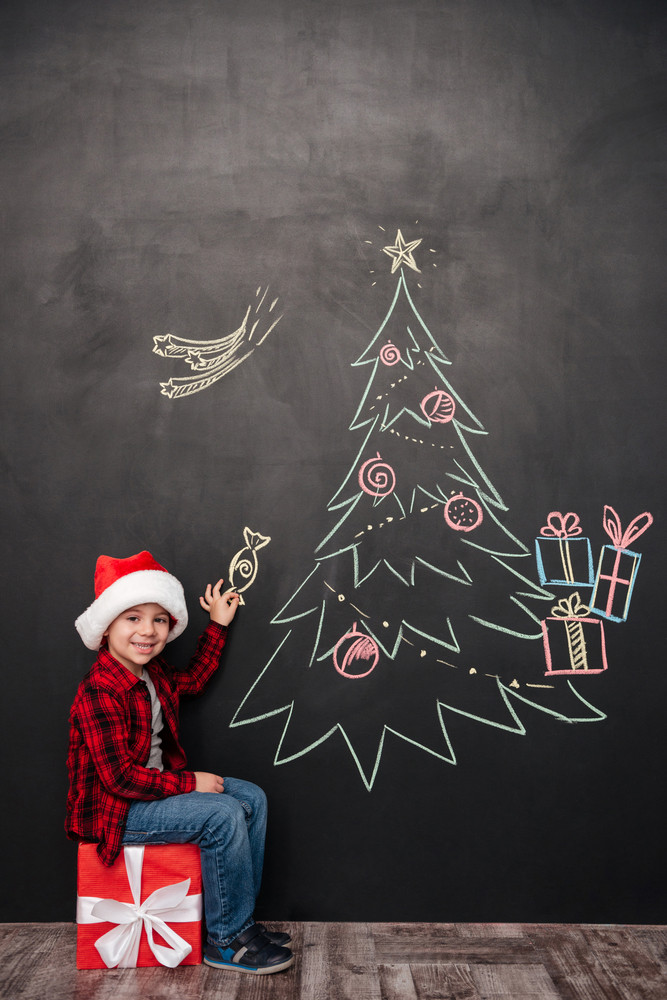 Image of happy child wearing hat sitting on big gift near Christmas tree drawing on blackboard while holding a candy. Looking at camera.