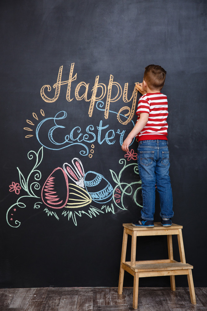 Little boy drawing easter doodles on chalk black board while standing on the ladder