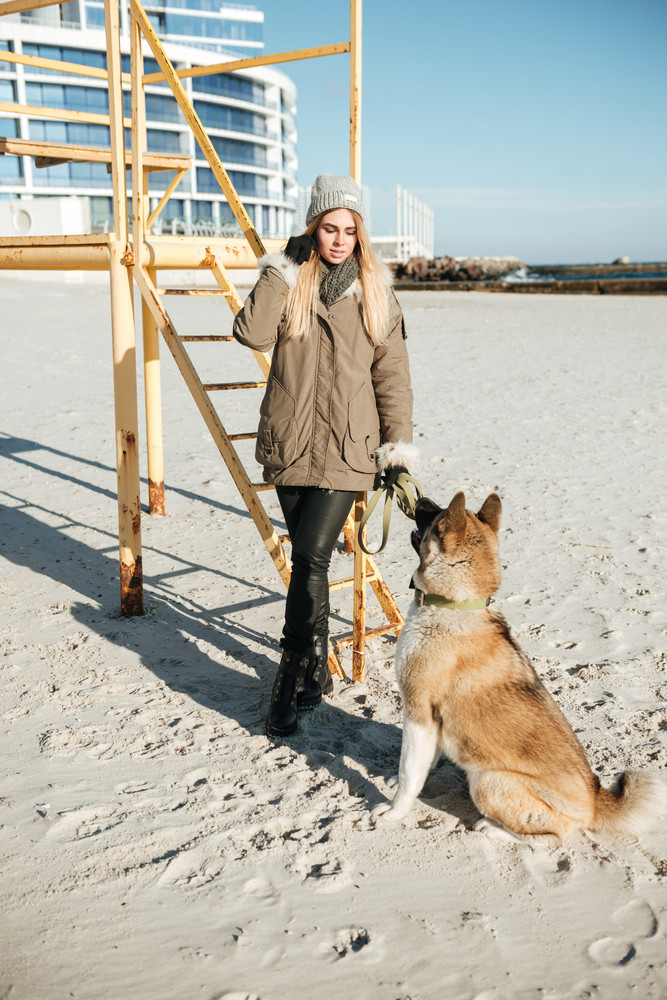 Photo of serious young woman walks in winter beach with dog on a leash.