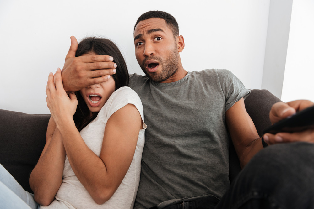 Picture of scared young couple watching TV on sofa at home. Man covering woman's eyes.