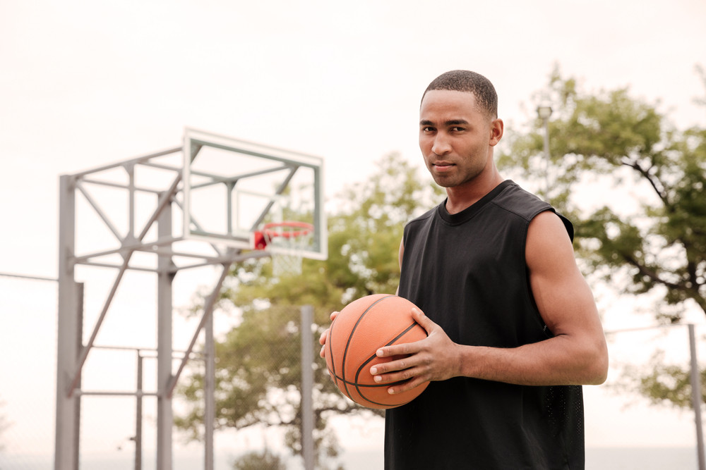 Portrait of african serious basketball player standing in the street with basketball hoop at background. Looking at camera.