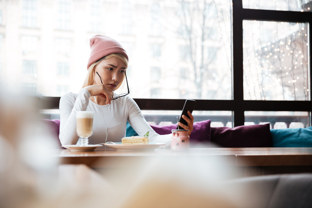 Sad frowning young woman in hat and glasses sitting and using mobile phone in cafe