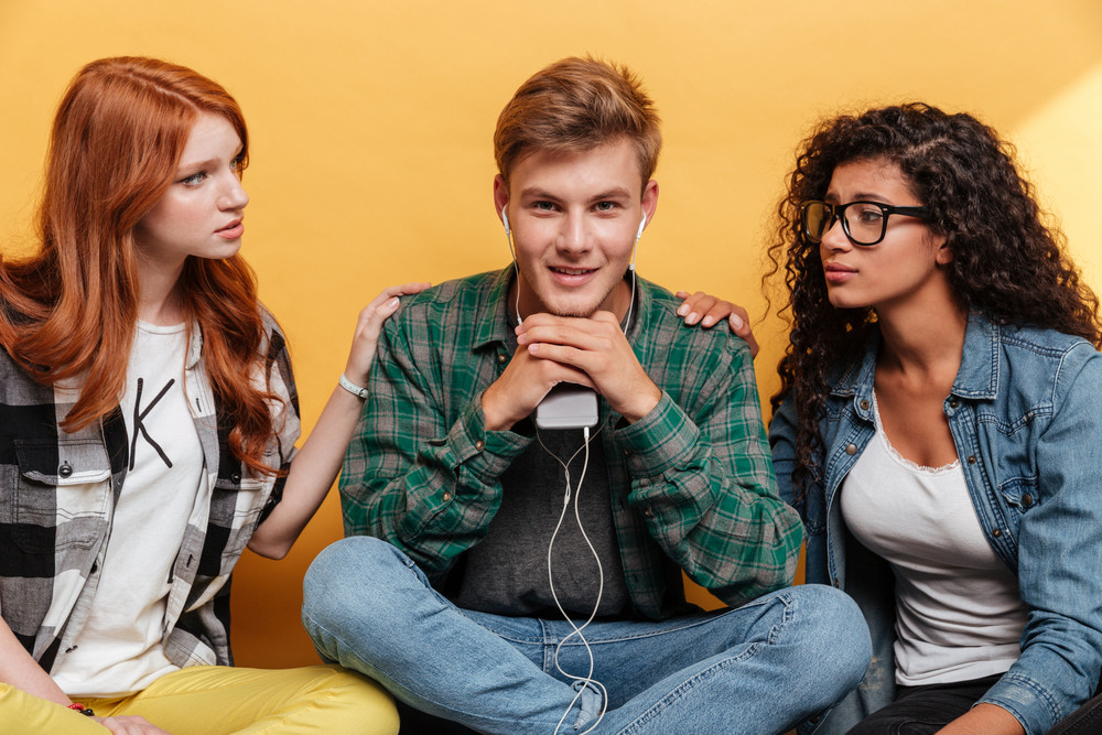 Two attractive young women fall in love in smiling man sitting and listening to music from cell phone over yellow background