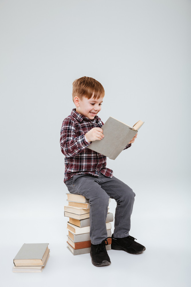 Vertical image of happy young boy in shirt reading book and sitting on books. Isolated gray background