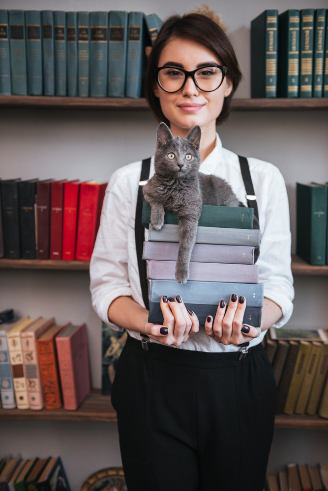 Vertical image of Smiling Authoress in glasses and white shirt holding books with cat on them and looking at camera
