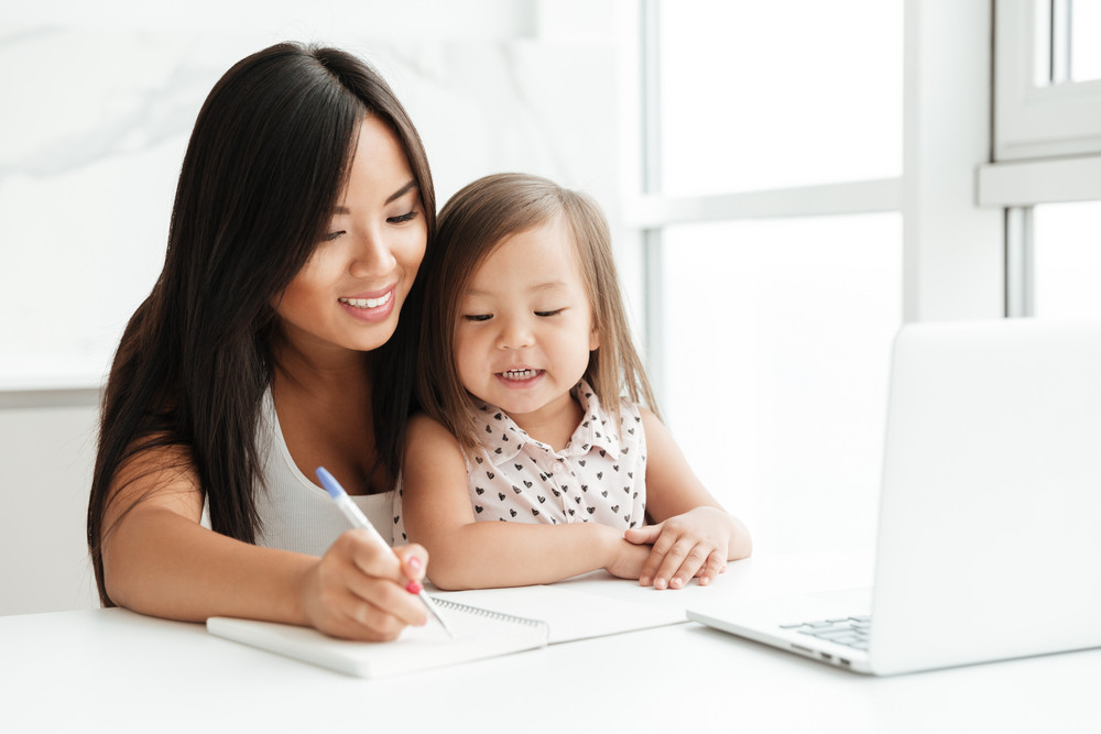 Image of amazing young mom sitting at the table with little cute asian girl at home indoors using laptop computer writing notes to notebook. Looking aside.