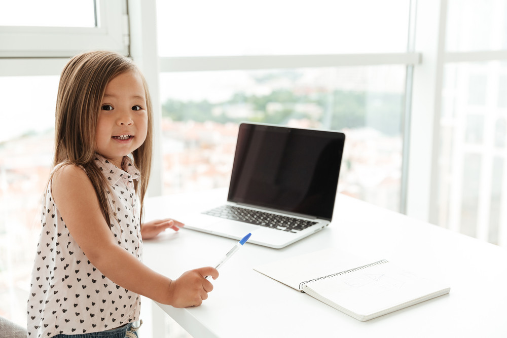 Little cute asian girl standing at home indoors near window using laptop computer and writing notes to notebook. Looking camera.