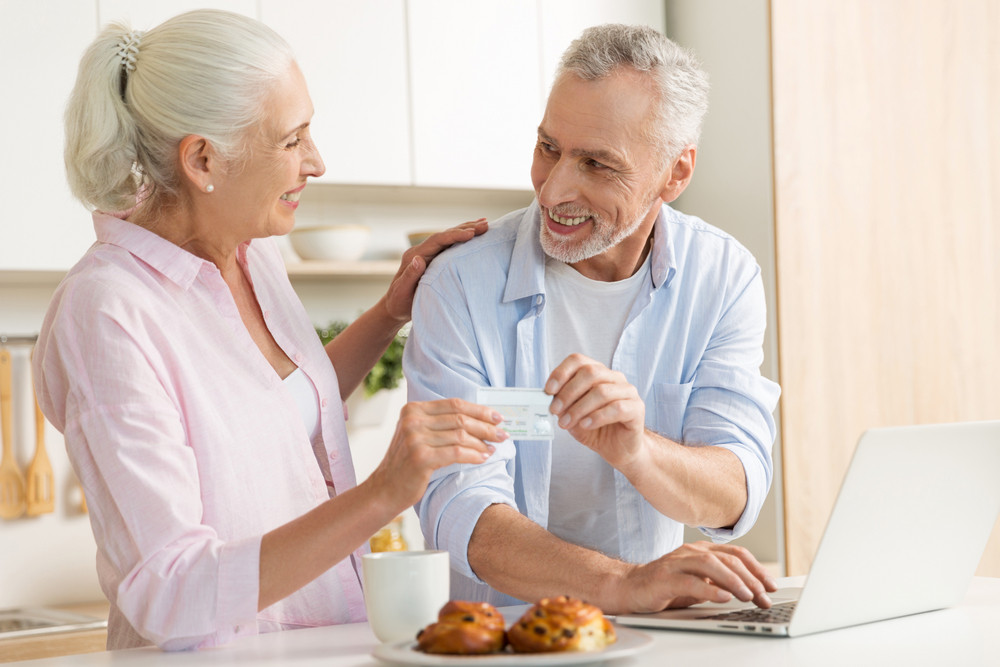 Picture of cheerful mature loving couple family standing at the kitchen using laptop computer holding credit card. Looking at each other.