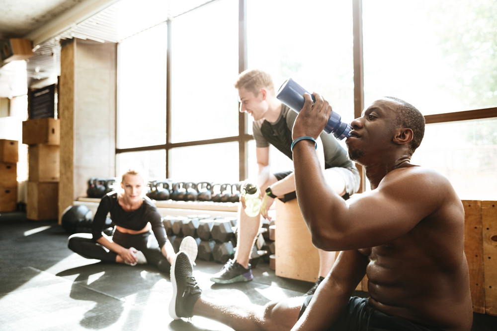 Picture of group of sports strong people sitting in gym. Looking aside drinking water.