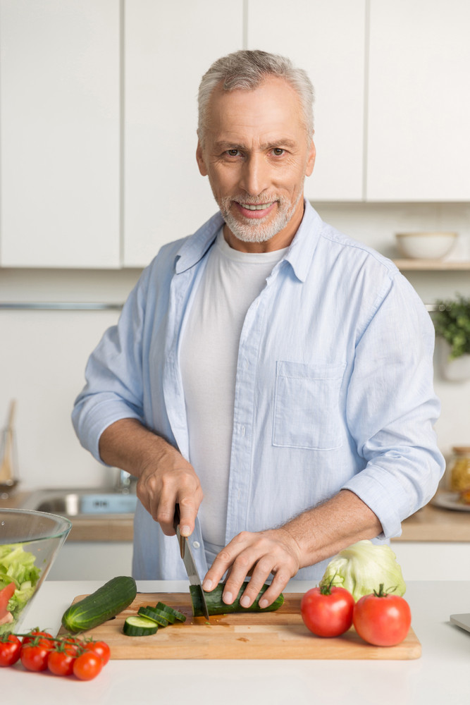 Picture of mature attractive man standing at the kitchen cooking salad. Looking at camera.