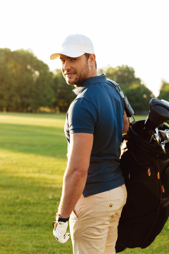 Smiling young man in cap holding golf bag and looking at camera while standing on a field