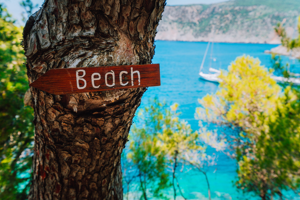 Assos village, Kefalonia. Greece. Beach wooden arrow sign on a pine tree showing direction to small hidden beach. White baot in sea background