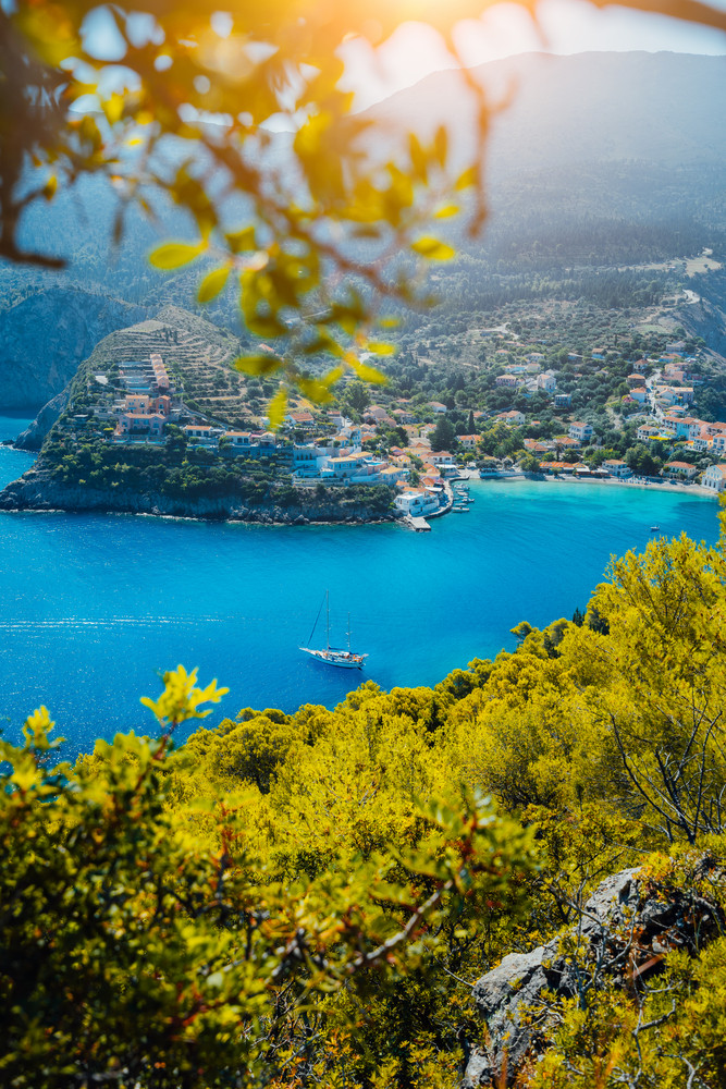 Assos village, Kefalonia. Greece. White yacht in blue bay framed by nature. Turquoise colored bay in Mediterranean sea surrounded by pine trees under bright sun beam light