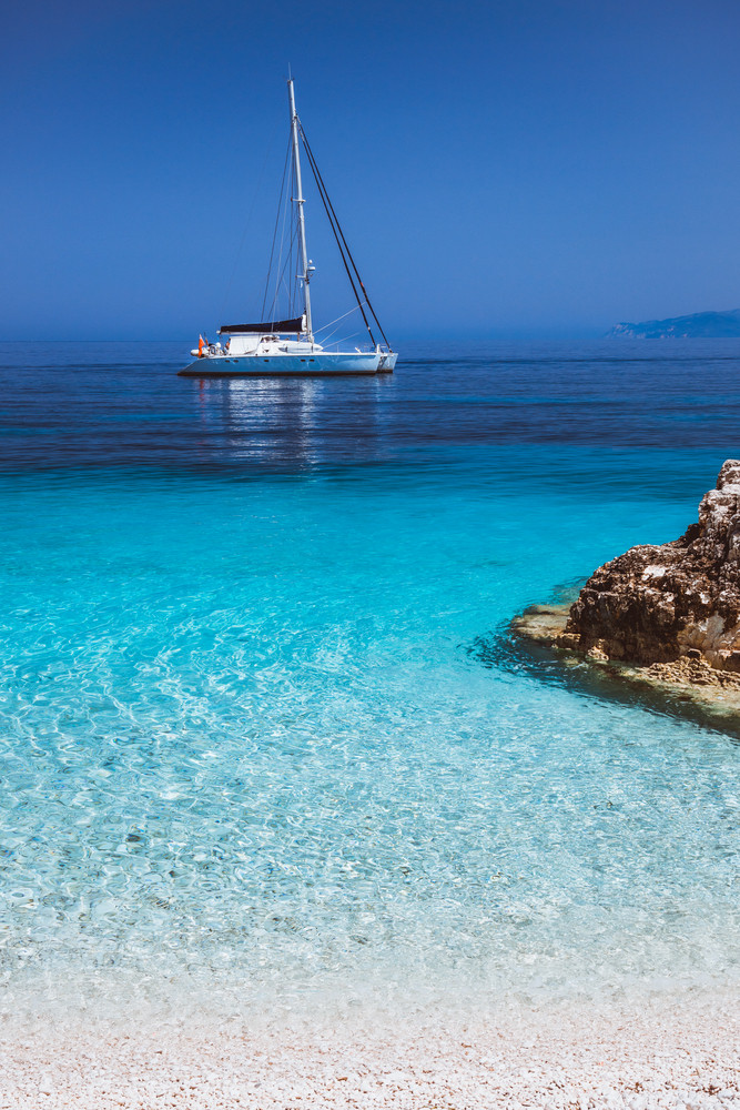 Beautiful calm azure blue lagoon with sailing catamaran yacht boat at anchor. Pure white pebble beach with rocks in the sea