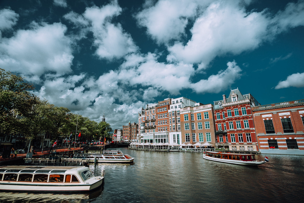 Classical Amsterdam cityscape. Cruise boats floating on the channel, river side promenade, cafes, typical Dutch architecture. Urban scene and white fluffy clouds