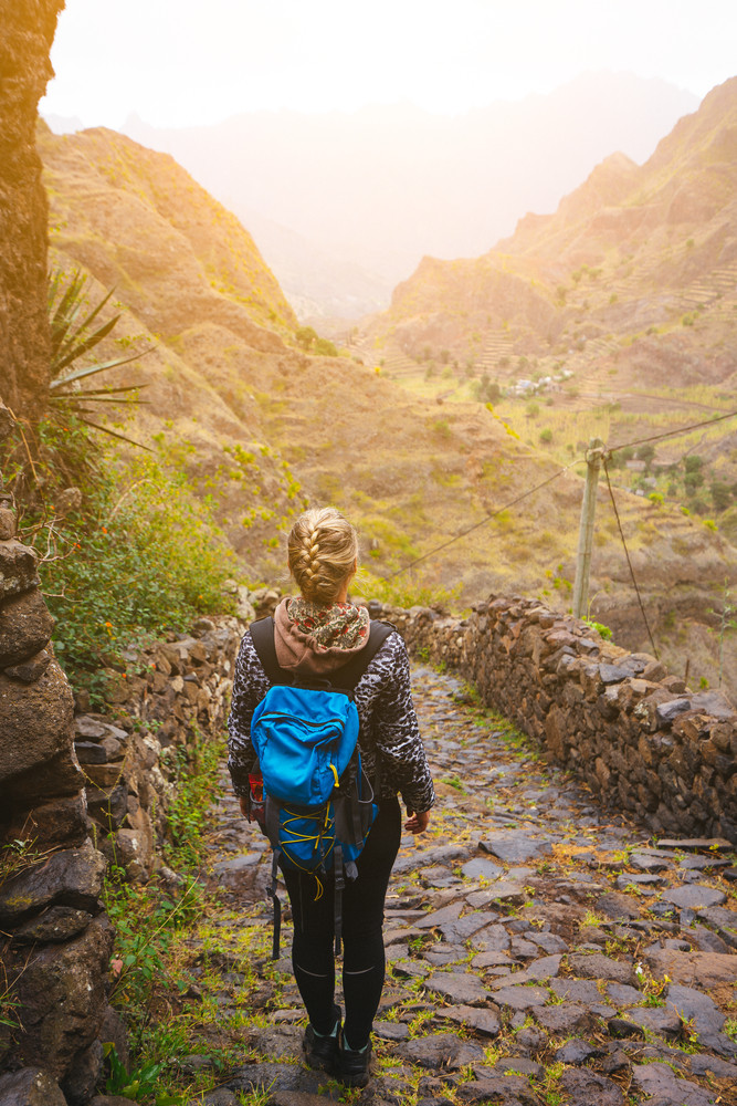 Female hiker with blue backpack staying on the stony cobbled trekking trail leading down to the valley between barren mountain peak on Santo Antao island, Cabo Verde